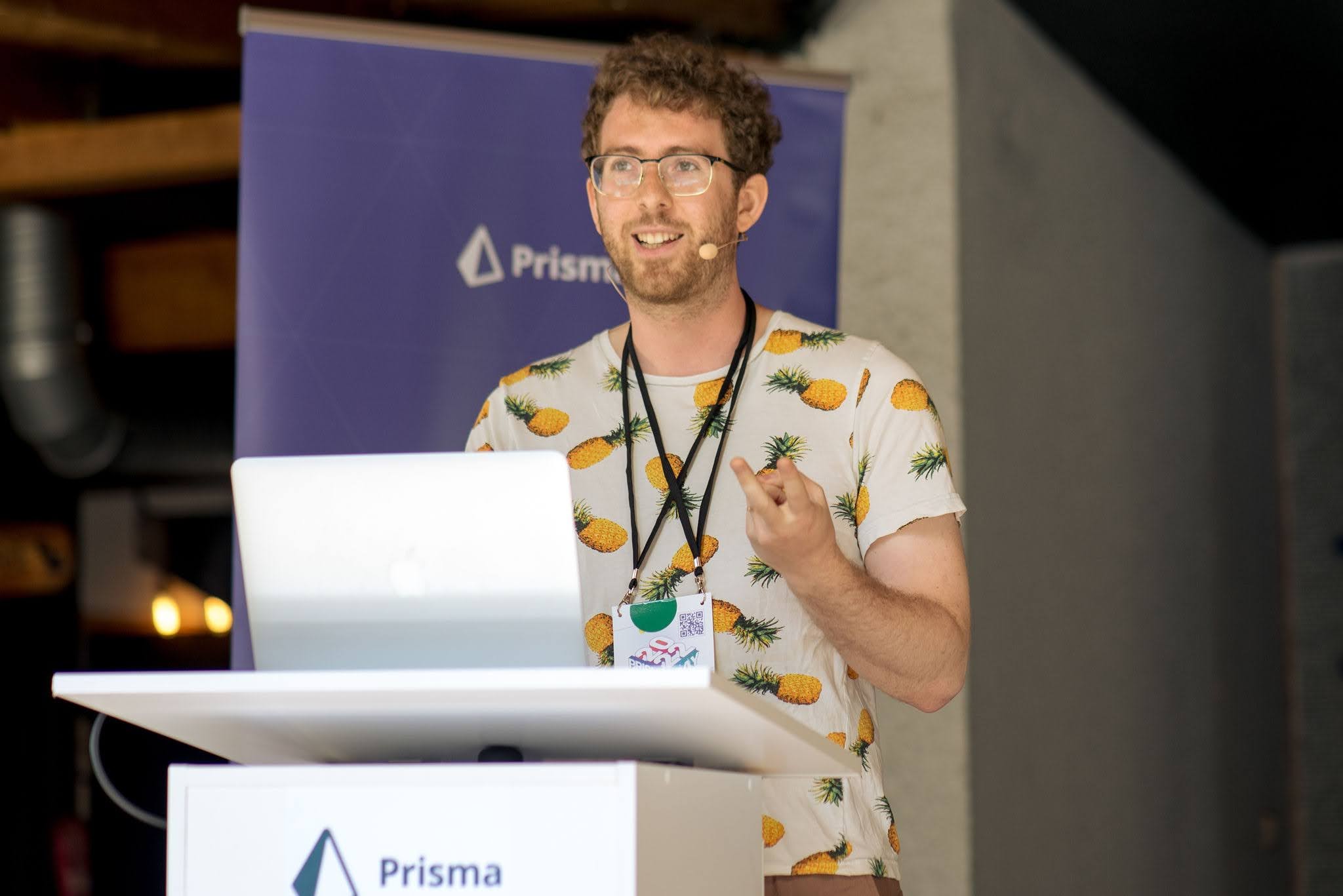 Me smiling and speaking at a conference podium, wearing a pineapples-on-white t-shirt, gesturing with one hand