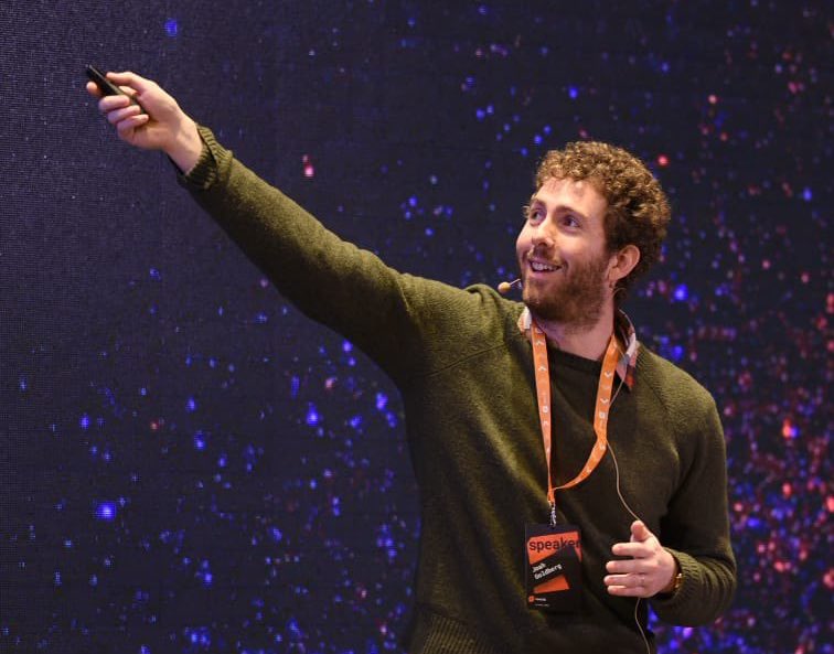 View from of me on stage, wearing a green sweater on a green, white, and red flannel with an orange revo.js lanyard around my neck, smiling and pointing a clicker up and to the side